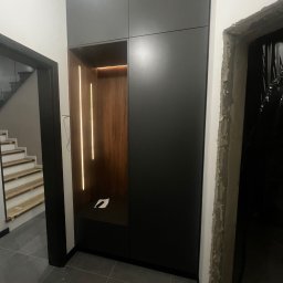 Stolarstwo - Ciemna szafa wnękowa z oświetleniem LED w przedpokoju, widok z perspektywy podłogi, surowe wykończenie ościeżnicy drzwi po prawej stronie.