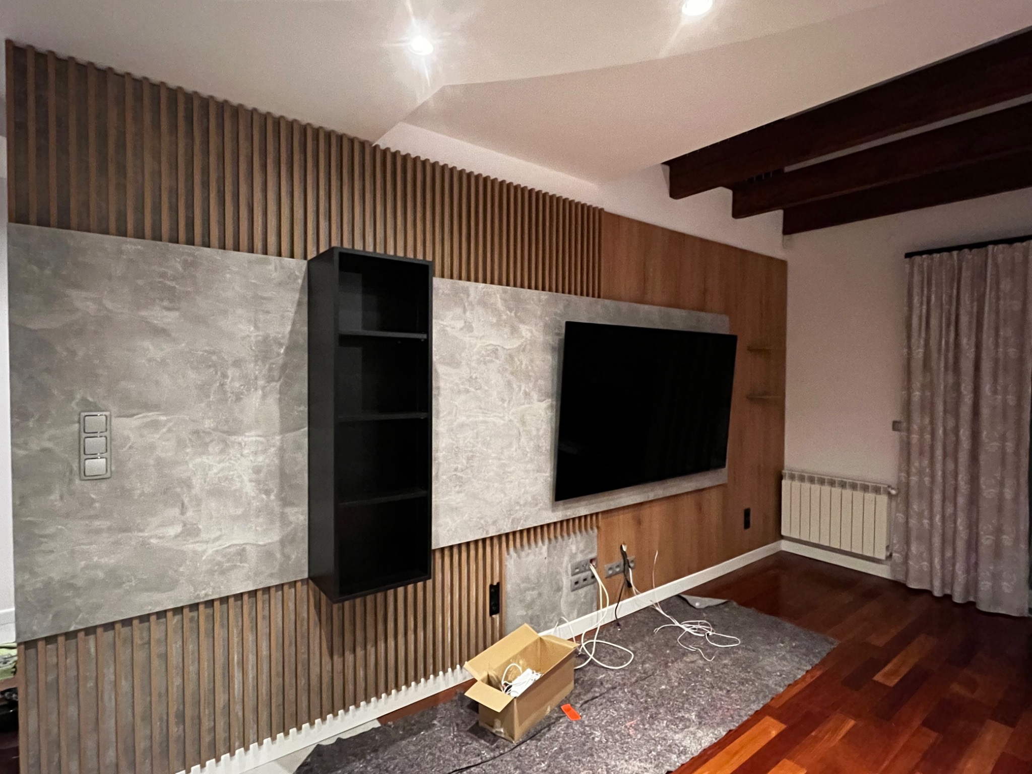 Nowoczesna ściana TV z drewnianymi lamelami, szarą płytą i czarną półką wiszącą. Widoczne kable i karton sugerują etap montażu.
