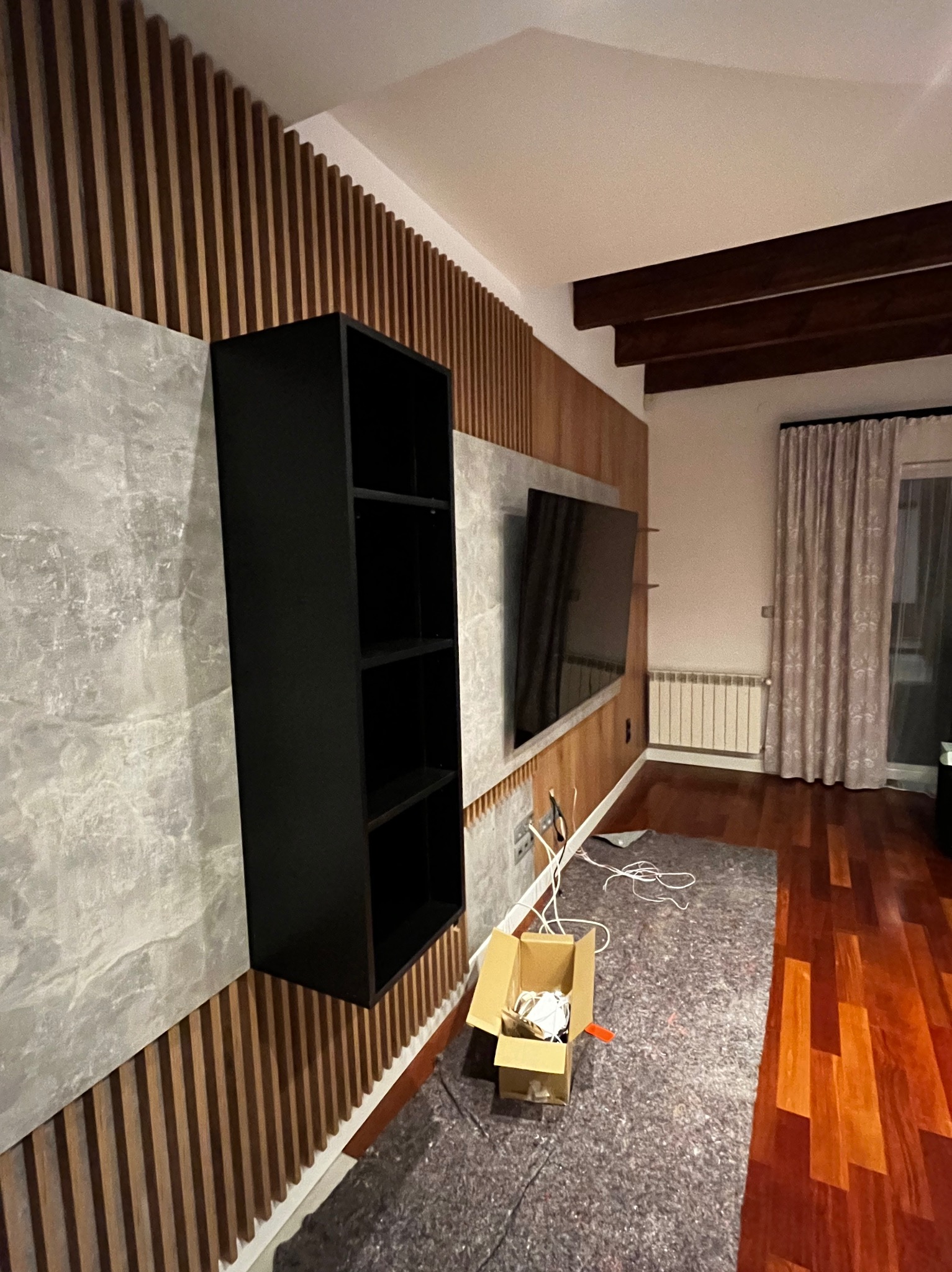 Czarna, wisząca szafka z półkami na ścianie z drewnianymi lamelami i szarą płytą imitującą beton. Na podłodze karton z akcesoriami montażowymi.