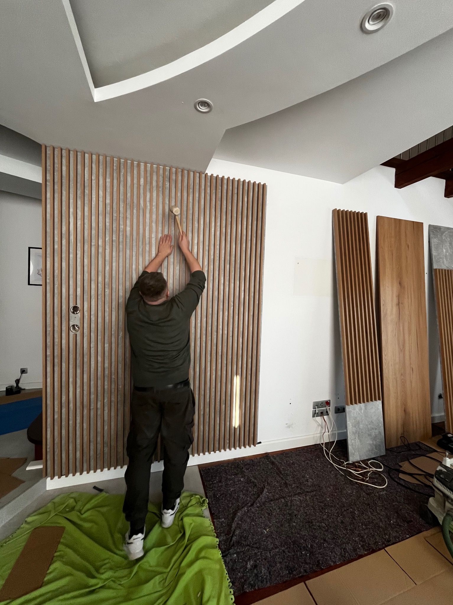 Montaż dekoracyjnych paneli ściennych z drewnianych listew. Widoczny pracownik w trakcie pracy, używający młotka. Częściowo widoczne panele czekają na montaż.