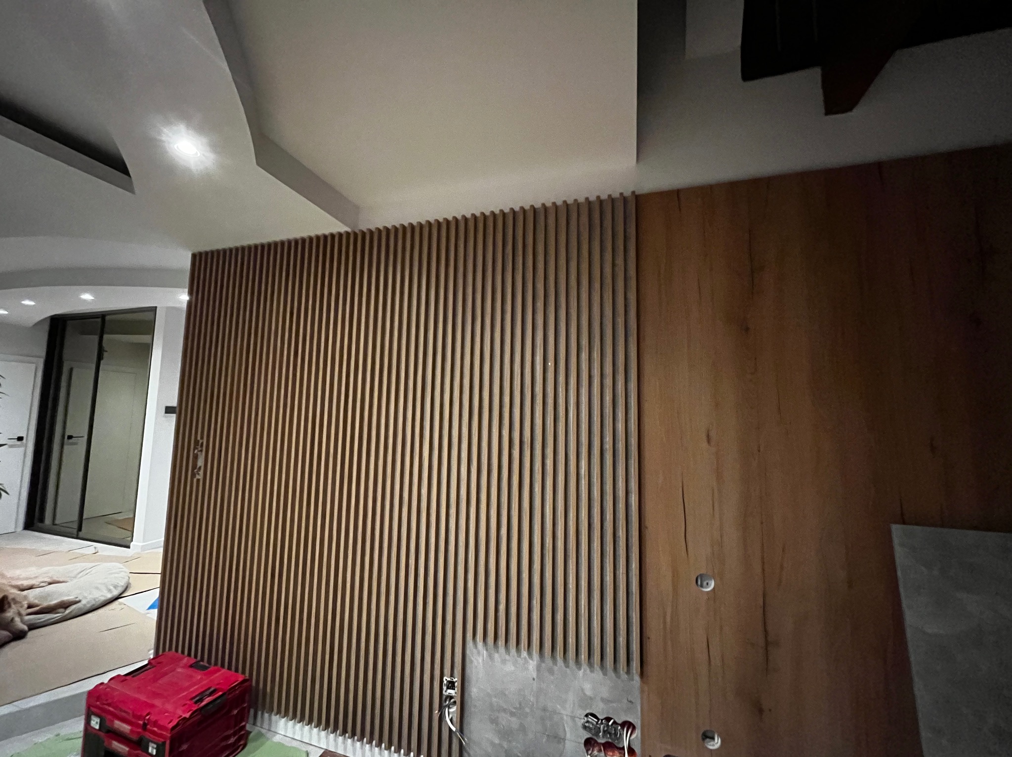 Drewniana ściana z pionowych listew i paneli w odcieniach brązu, z widocznymi elementami instalacji elektrycznej w trakcie montażu. W tle widoczne pomieszczenie.