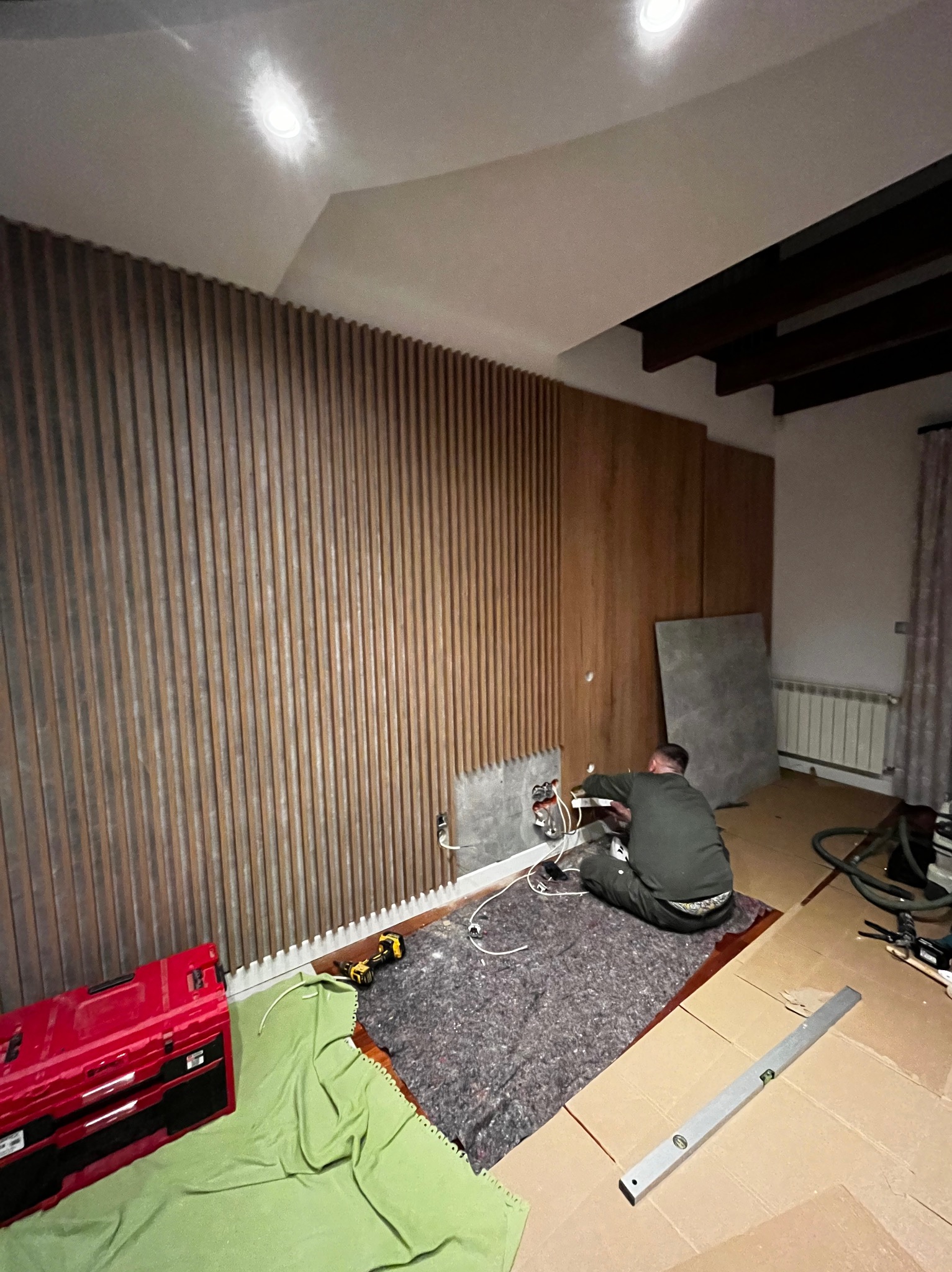 Montaż drewnianej ściany dekoracyjnej z pionowych listew. Elektryk pracuje nad instalacją elektryczną. Widoczna skrzynka narzędziowa i poziomica.