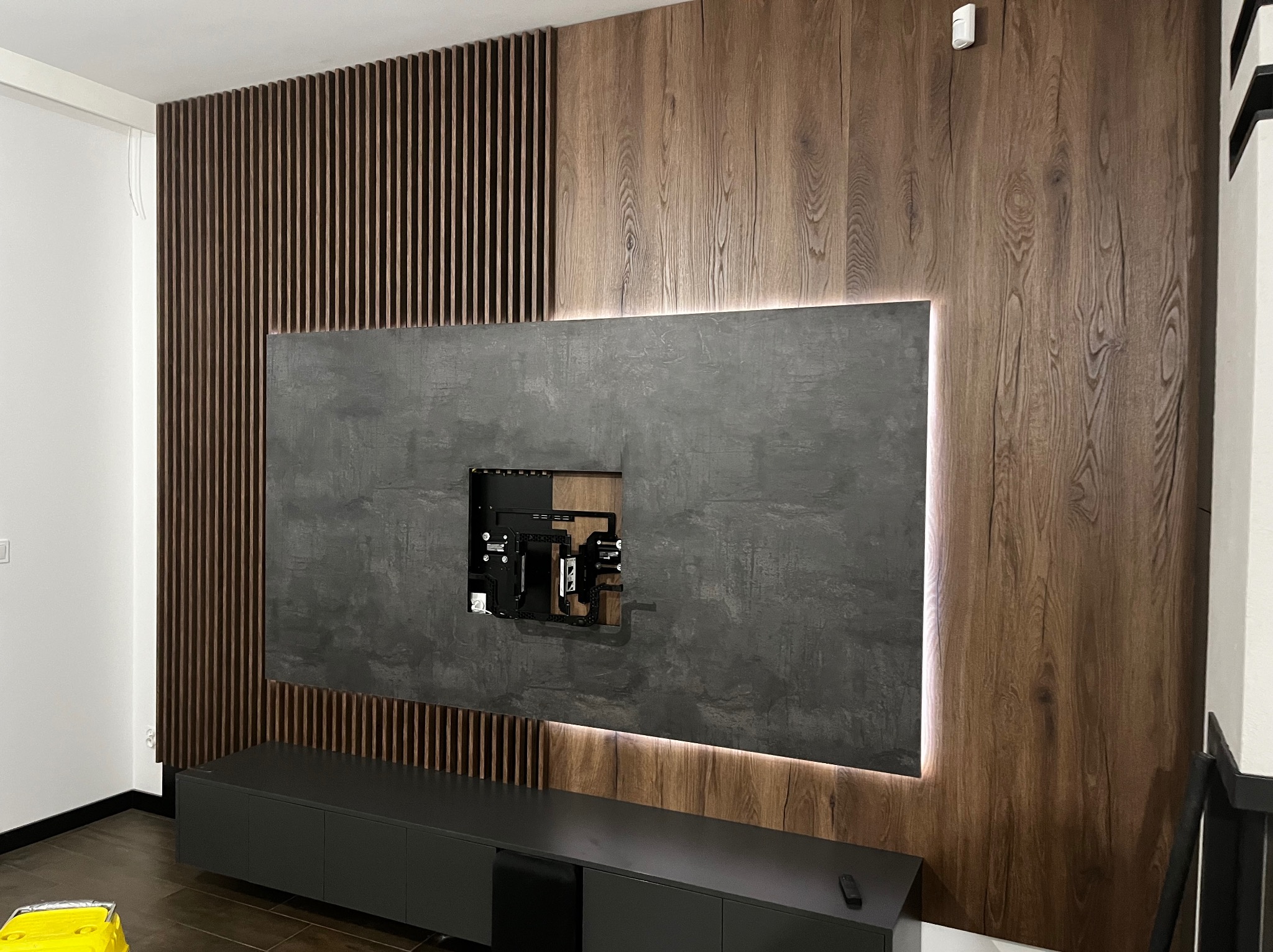 Nowoczesna ściana TV z drewnianymi lamelami, szarą płytą i podświetleniem LED. Minimalistyczna szafka RTV w kolorze grafitowym. Widoczne mocowanie telewizora.