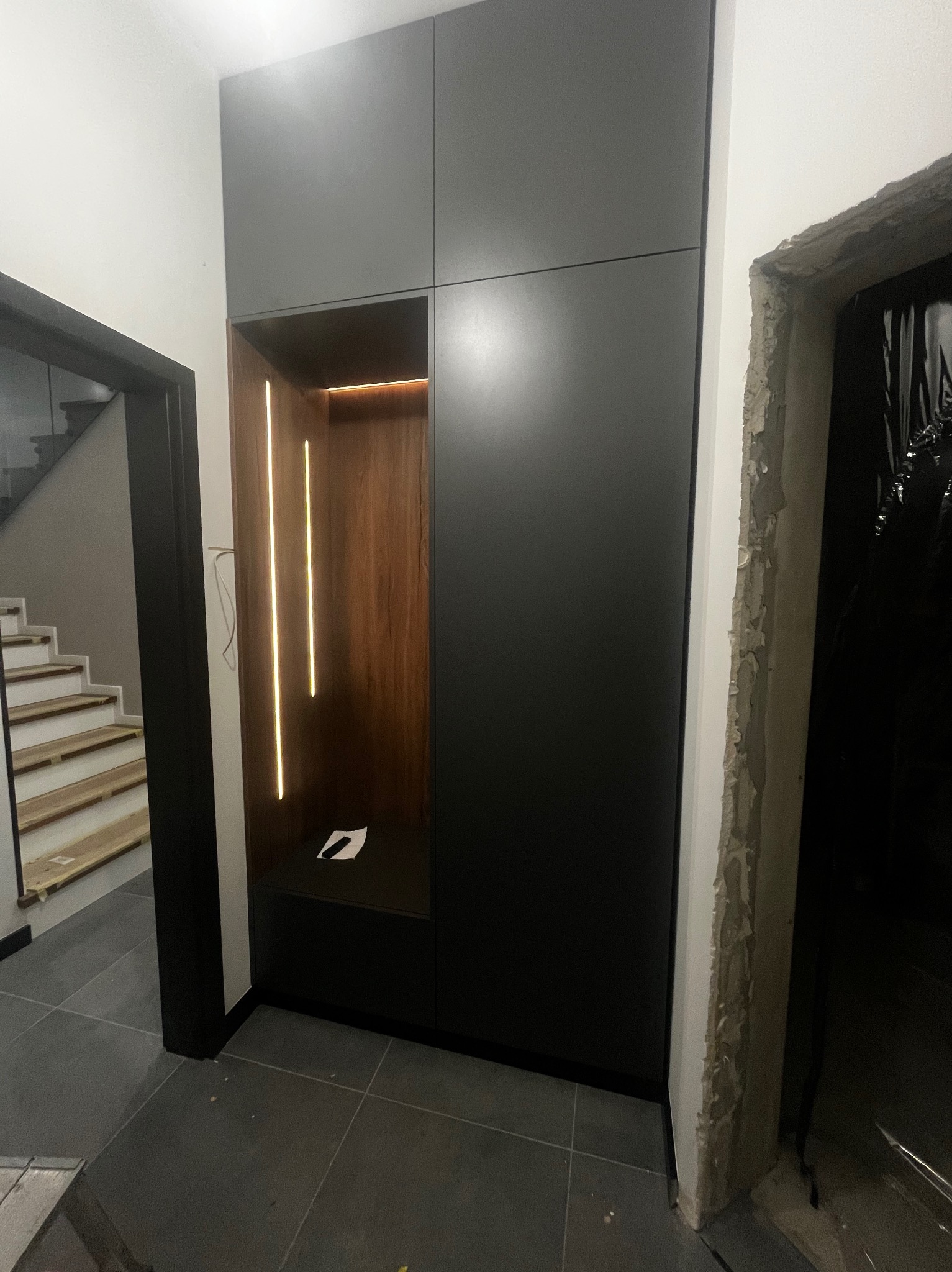 Ciemna szafa wnękowa z oświetleniem LED w przedpokoju, widok z perspektywy podłogi, surowe wykończenie ościeżnicy drzwi po prawej stronie.