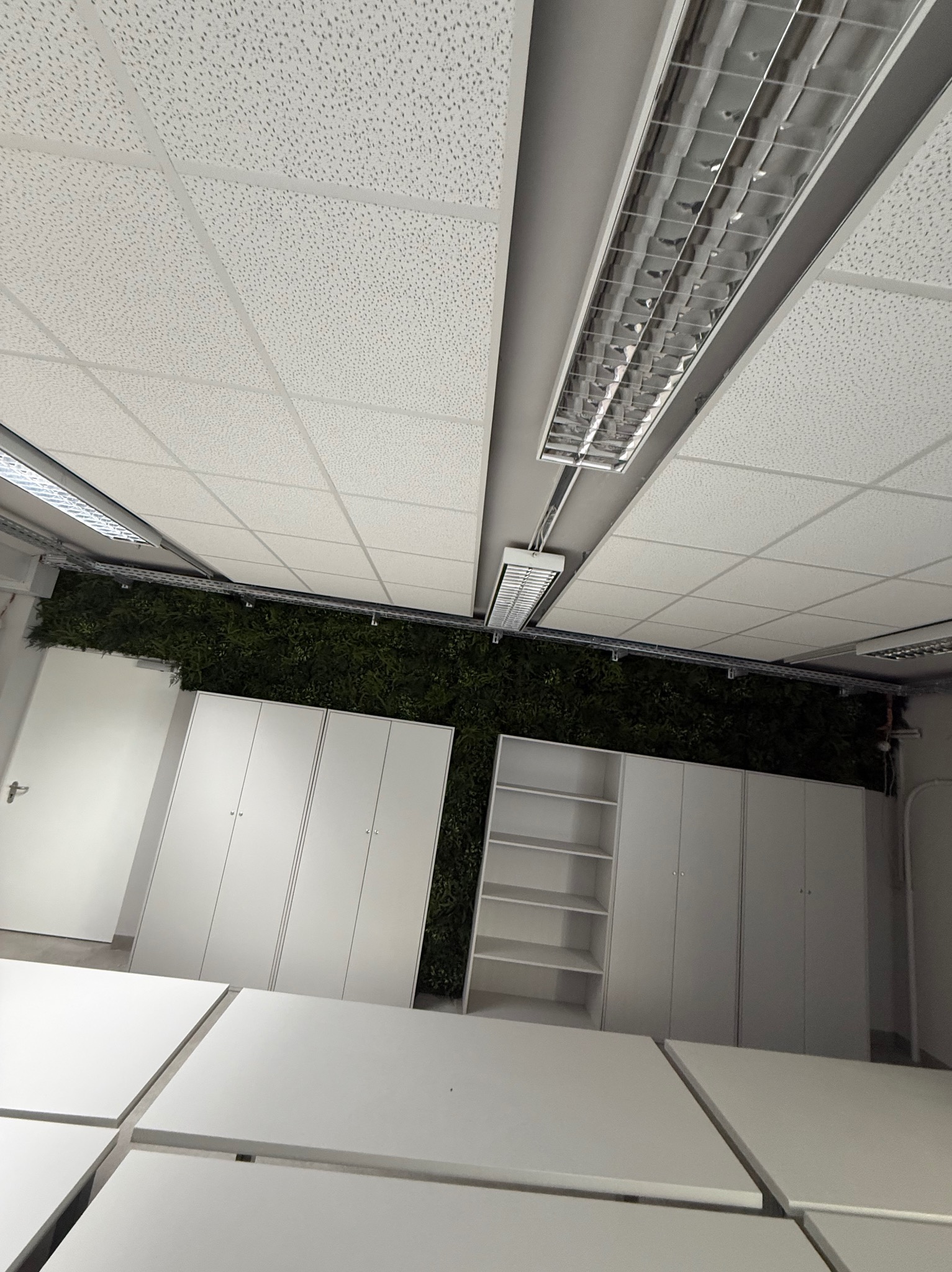 Białe szafy z zieloną ścianą w tle, oświetlone lampami sufitowymi. Widok z dołu, ukazujący nowoczesne meble i elementy dekoracyjne.