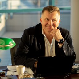 SZAŁAŃSKI I PARTNERZY KANCELARIA RESTRUKTURYZACYJNA SPÓŁKA Z OGRANICZONĄ ODPOWIEDZIALNOŚCIĄ - Portret mężczyzny w biurze, z laptopem i zieloną lampką. Uśmiechnięty, z zegarkiem na ręku, oparty o dłoń. W tle okno i kolumna.