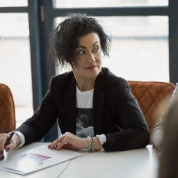 SZAŁAŃSKI I PARTNERZY KANCELARIA RESTRUKTURYZACYJNA SPÓŁKA Z OGRANICZONĄ ODPOWIEDZIALNOŚCIĄ - Kobieta w czarnej marynarce, siedząca przy stole z dokumentem Fenix, podczas spotkania z innymi osobami w jasnym biurze z dużymi oknami.