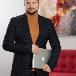 SZAŁAŃSKI I PARTNERZY KANCELARIA RESTRUKTURYZACYJNA SPÓŁKA Z OGRANICZONĄ ODPOWIEDZIALNOŚCIĄ - Mężczyzna w marynarce w kratę i golfie trzyma laptopa Apple na tle czerwonej sofy i abstrakcyjnego obrazu. Profesjonalny wizerunek prawnika.