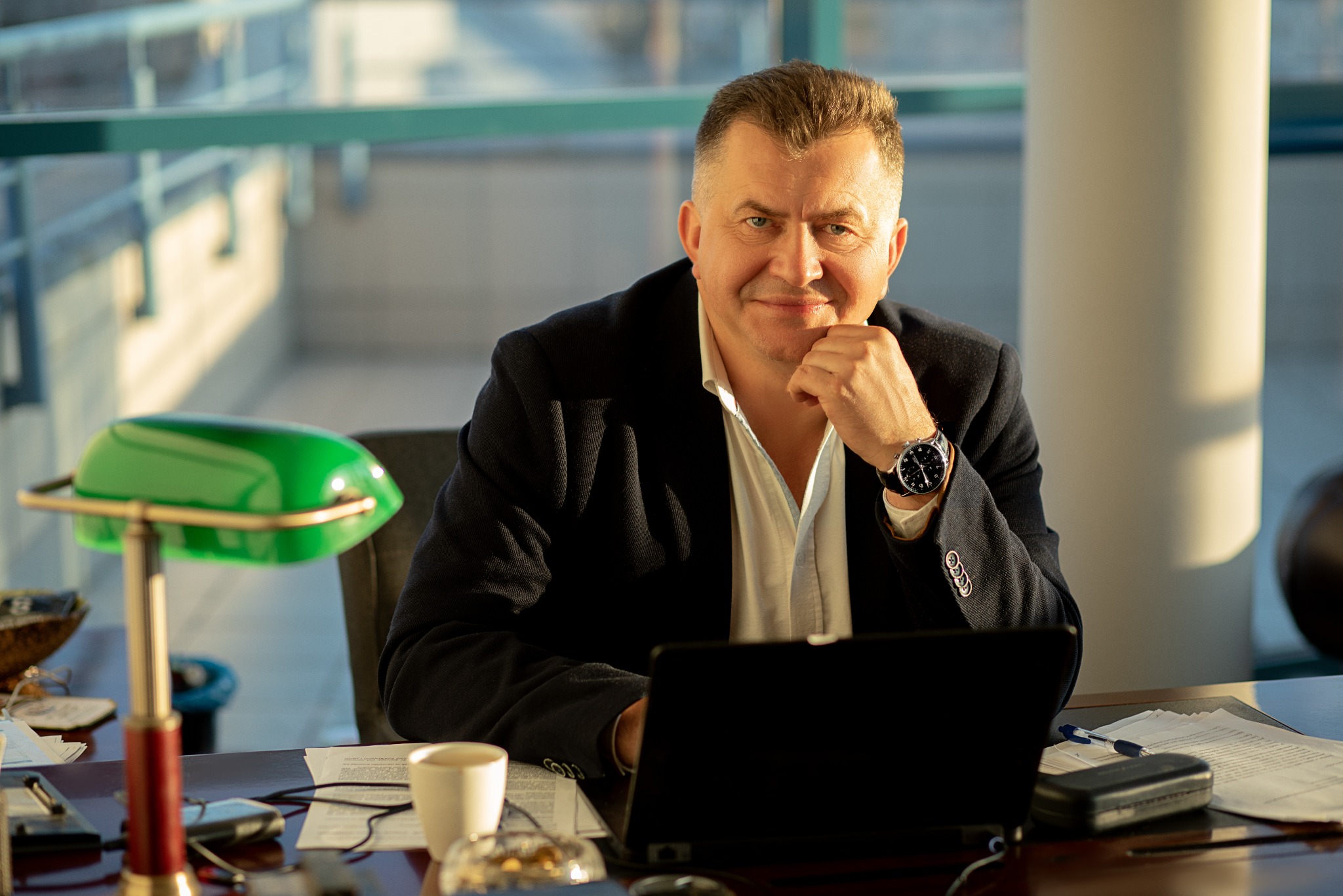 Portret mężczyzny w biurze, z laptopem i zieloną lampką. Uśmiechnięty, z zegarkiem na ręku, oparty o dłoń. W tle okno i kolumna.