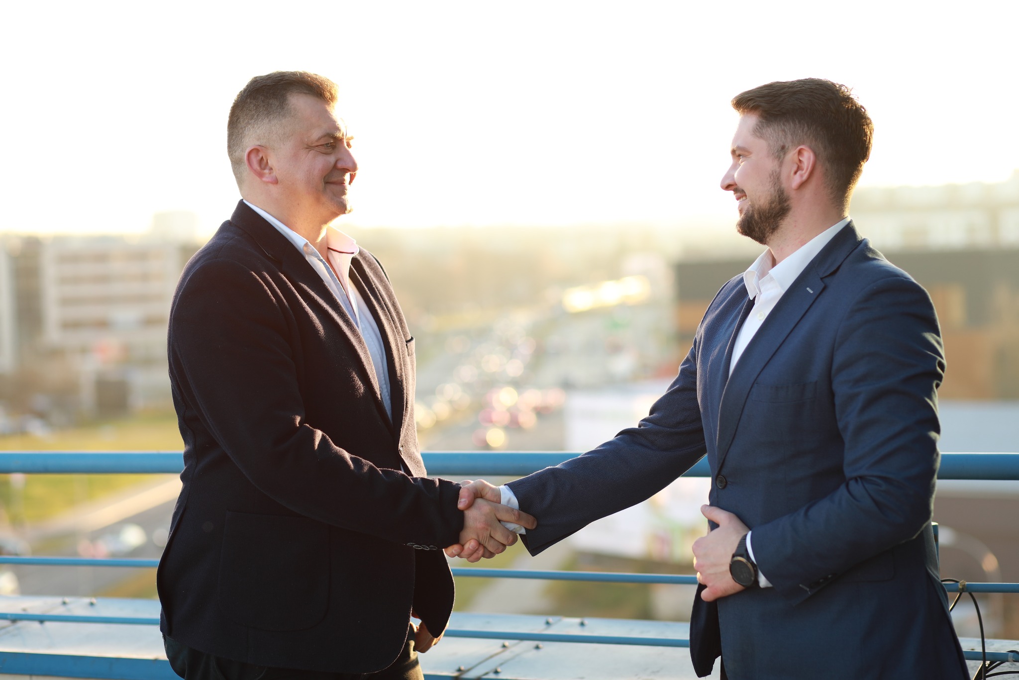 Uścisk dłoni dwóch mężczyzn w garniturach na tle panoramy Warszawy. Symbolizuje zawarcie umowy, partnerstwo biznesowe, zaufanie i profesjonalizm.