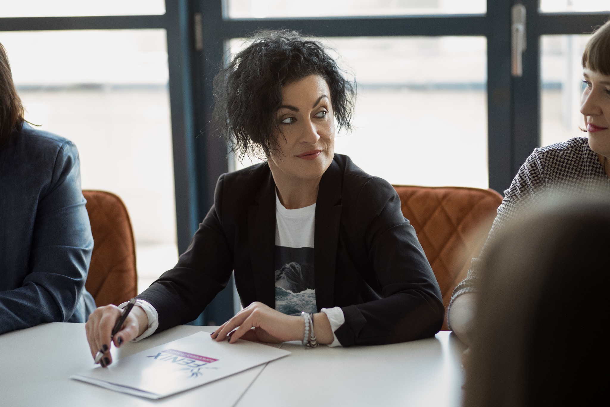 Kobieta w czarnej marynarce, siedząca przy stole z dokumentem Fenix, podczas spotkania z innymi osobami w jasnym biurze z dużymi oknami.