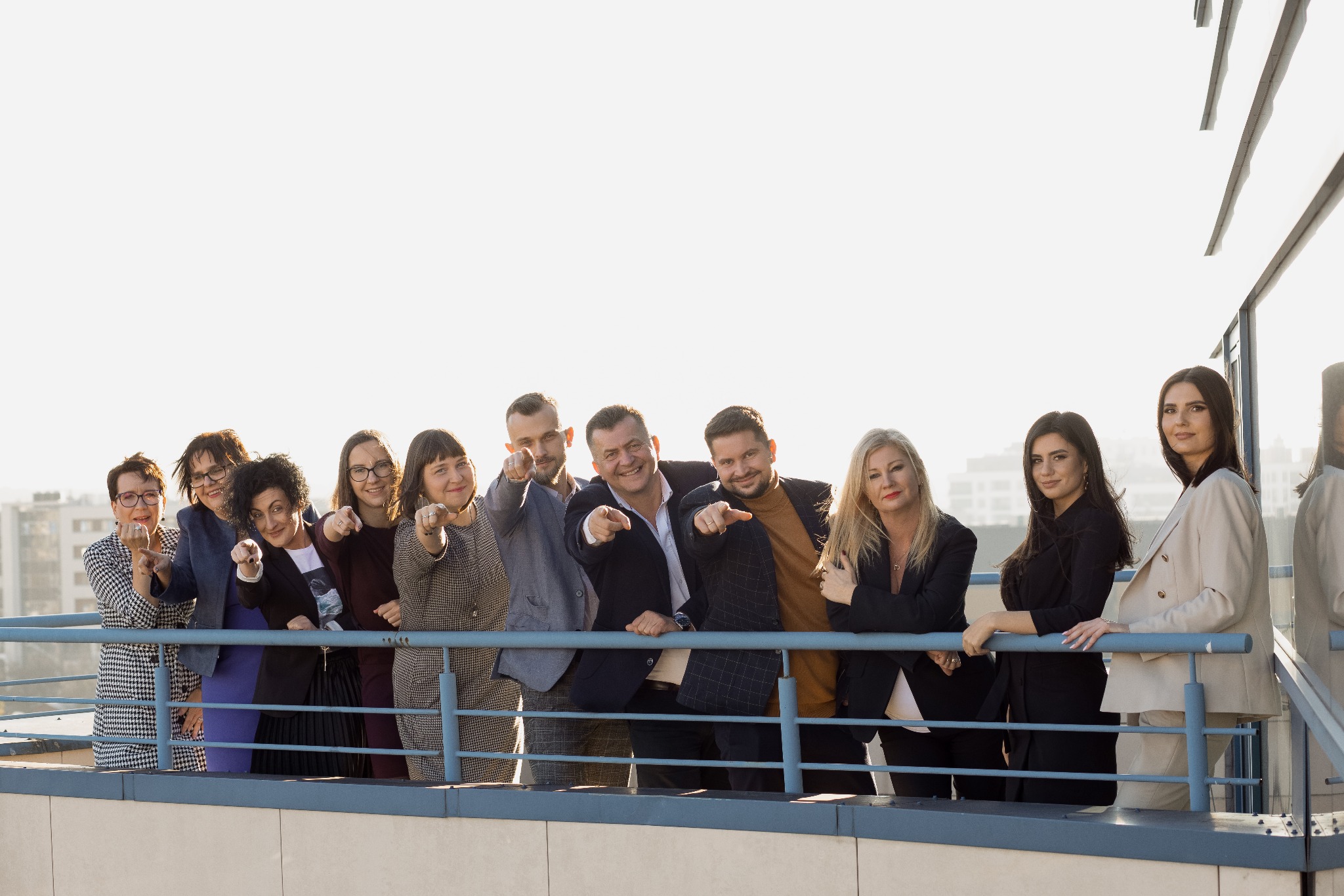 Zespół prawników na tle miasta, wskazujący palcami w stronę widza. Uśmiechnięci, w biznesowych strojach, stoją na balkonie z metalową barierką.