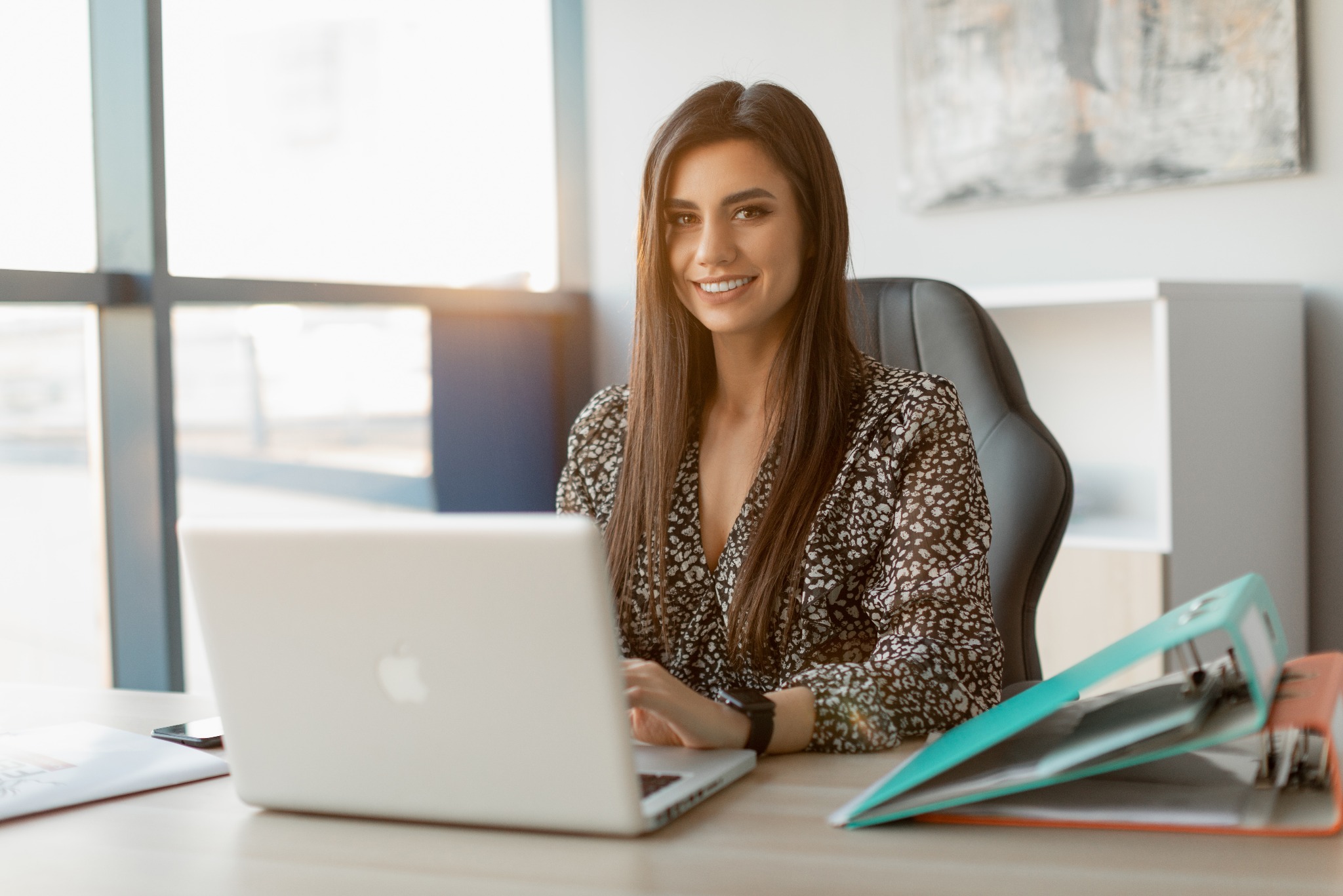 Uśmiechnięta brunetka w biurze, siedząca przy biurku z laptopem i segregatorami, w tle okno i regał. Delikatne światło słoneczne wpada do pomieszczenia.
