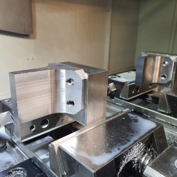 Sop-Tech Tomasz Sobczak - Obrabiany, metalowy element z dwoma otworami, zamocowany w uchwycie maszyny CNC. Widoczne wióry i chłodziwo. Precyzyjna obróbka skrawaniem.