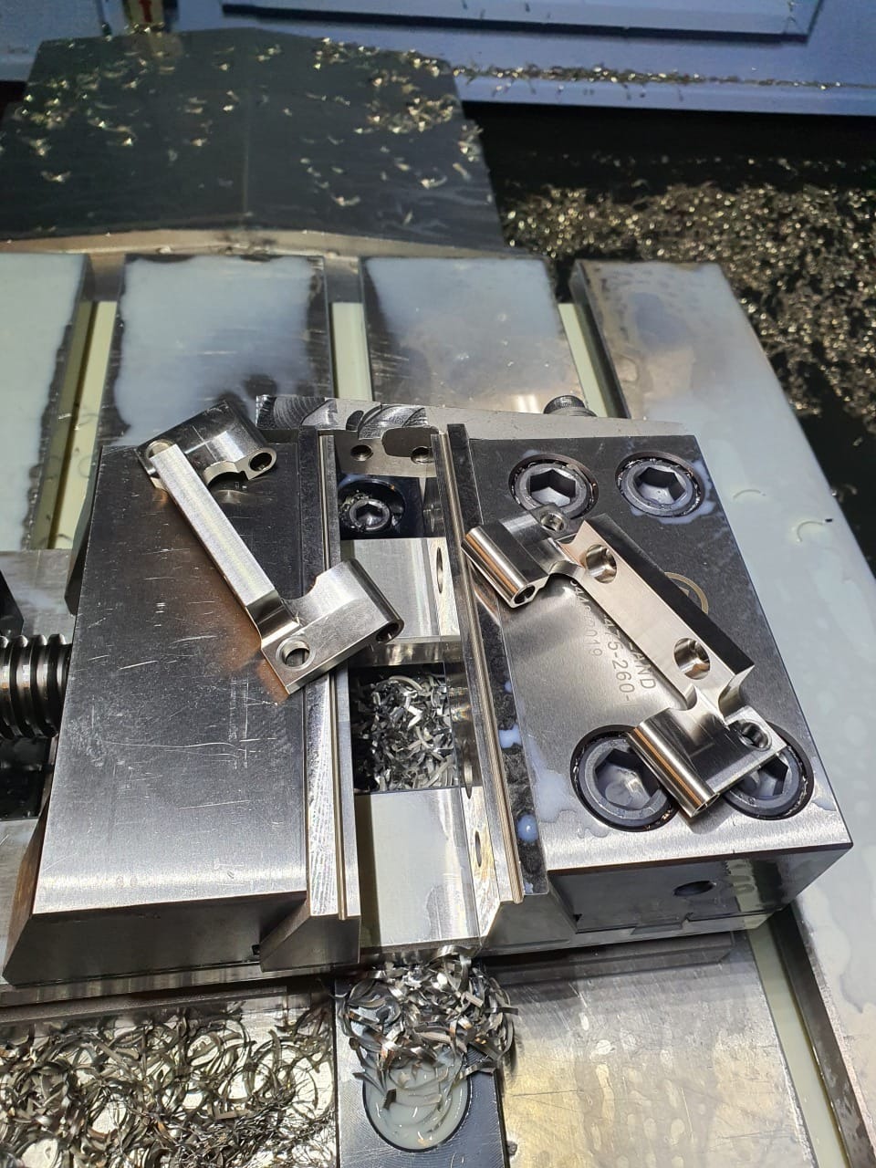 Metalowy uchwyt z obrobionymi elementami na tle wiórów i chłodziwa, detale po precyzyjnej obróbce, widoczne ślady frezowania, surowy przemysłowy klimat.