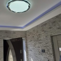 Mateusz Bielański Remonty i Wykończenia Wnętrz - Wykończone wnętrze z dekoracyjną ścianą z kamienia, oświetleniem LED na suficie i nowoczesną lampą. Widoczny fragment drzwi.