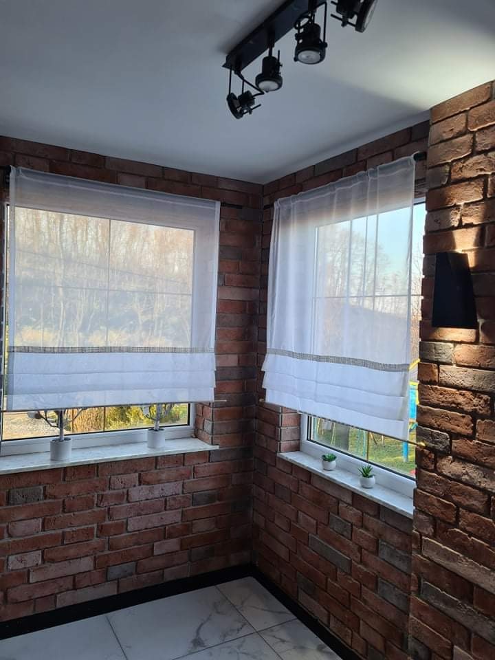 Balkon z ceglaną ścianą, dwoma oknami z białymi roletami rzymskimi i małymi roślinami na parapetach. Na suficie czarna lampa z kilkoma reflektorami.