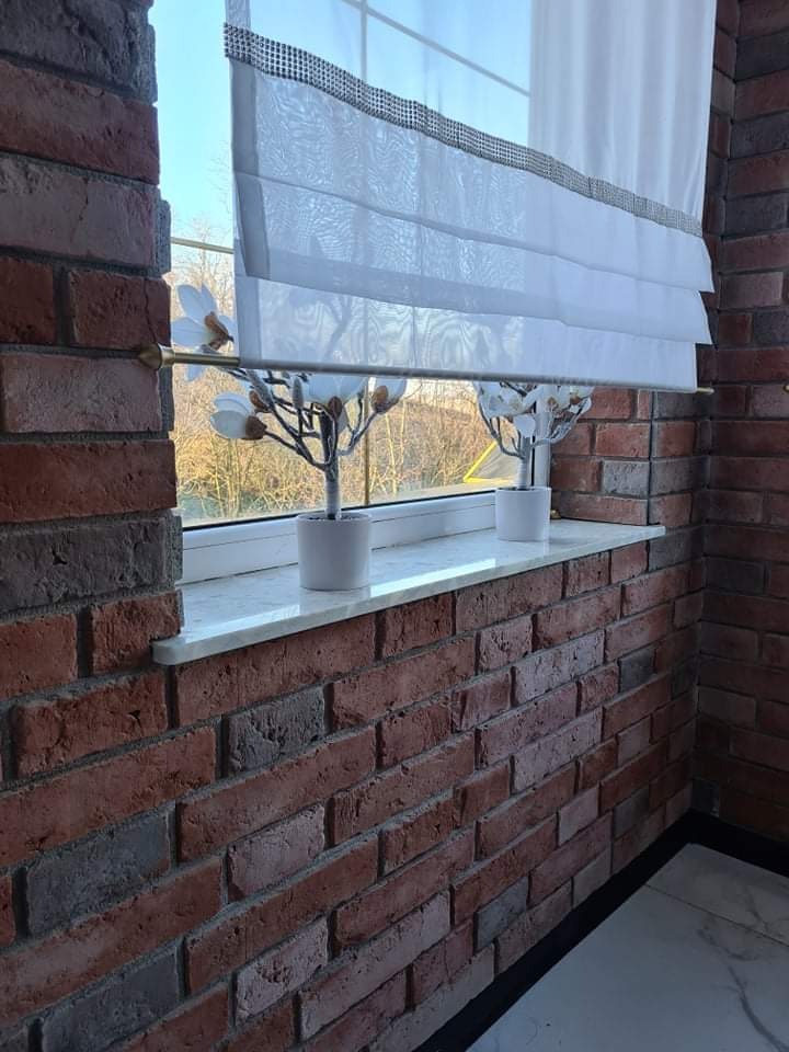 Balkon z ceglaną ścianą, białym parapetem i roletą rzymską. Dwa małe drzewka w doniczkach na parapecie. Widok z okna na zewnątrz. Dekoracyjne wykończenie.