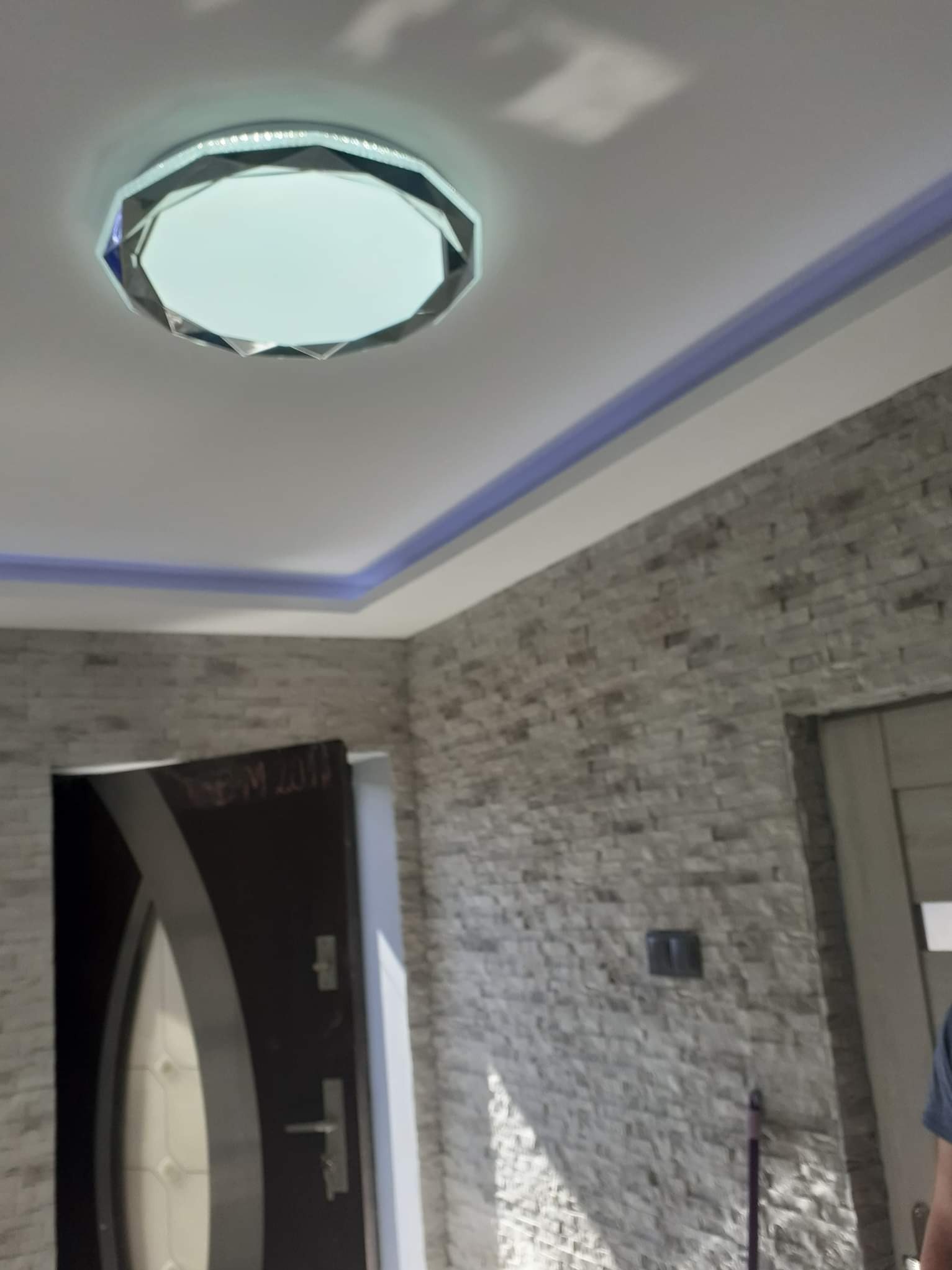 Wykończone wnętrze z dekoracyjną ścianą z kamienia, oświetleniem LED na suficie i nowoczesną lampą. Widoczny fragment drzwi.