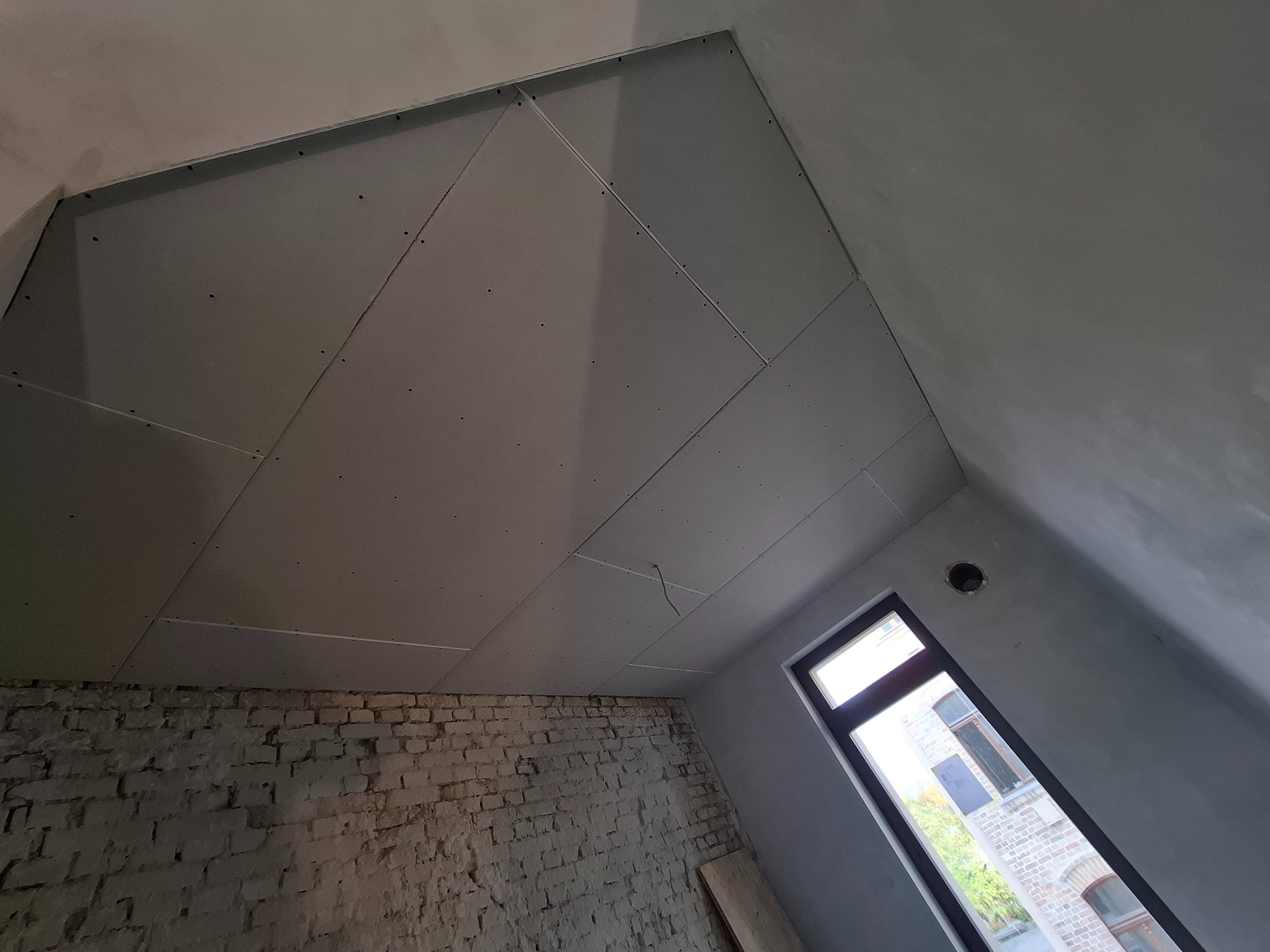 Skośny sufit z płyt karton-gips na poddaszu, widoczna ceglana ściana i okno. Prace wykończeniowe w trakcie, surowy stan.