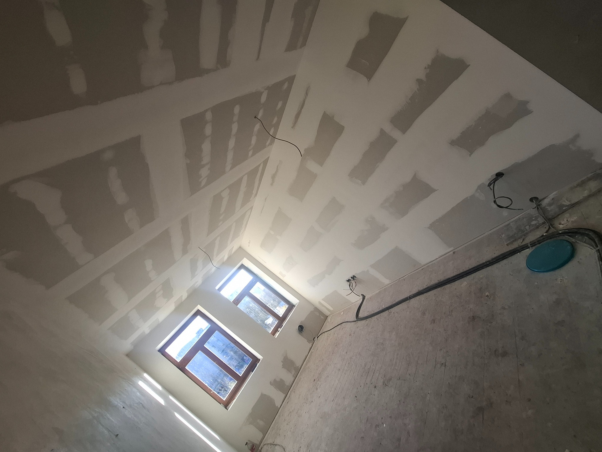 Skośny sufit poddasza z oknami, pokryty płytami kartonowo-gipsowymi, częściowo zaszpachlowanymi. Widoczne kable i surowe ściany. Ujęcie z dołu.