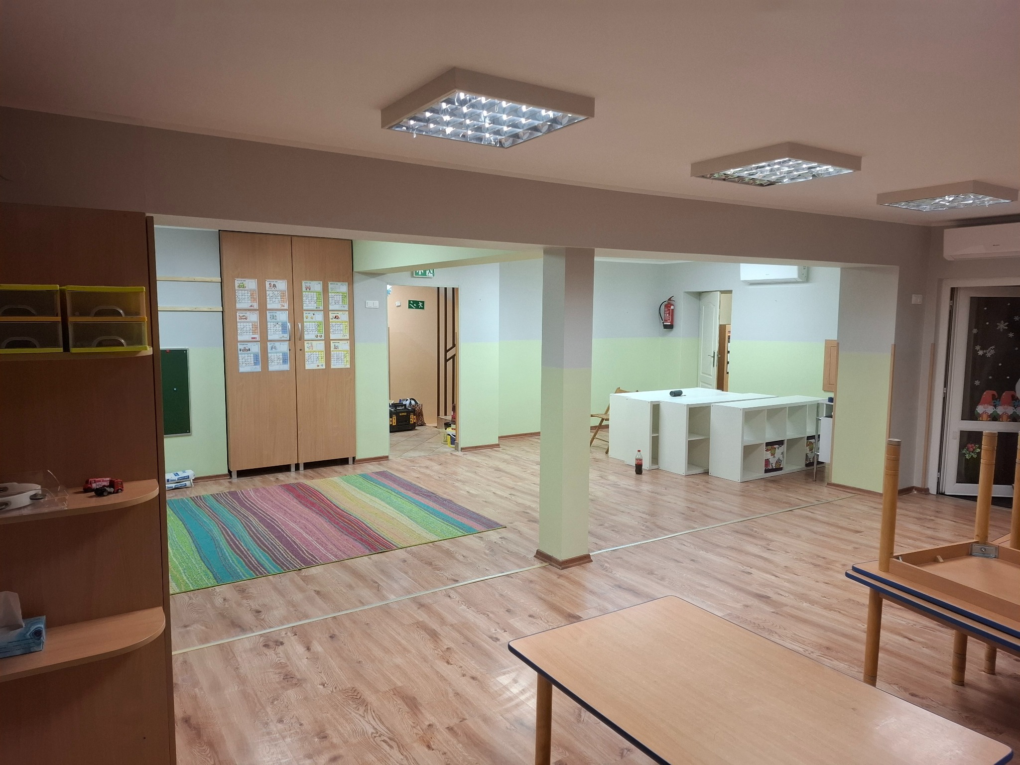 Jasne wnętrze sali przedszkolnej po remoncie: drewniana podłoga, kolorowy dywan, meble i oświetlenie. Widoczne detale wykończenia i aranżacji przestrzeni.