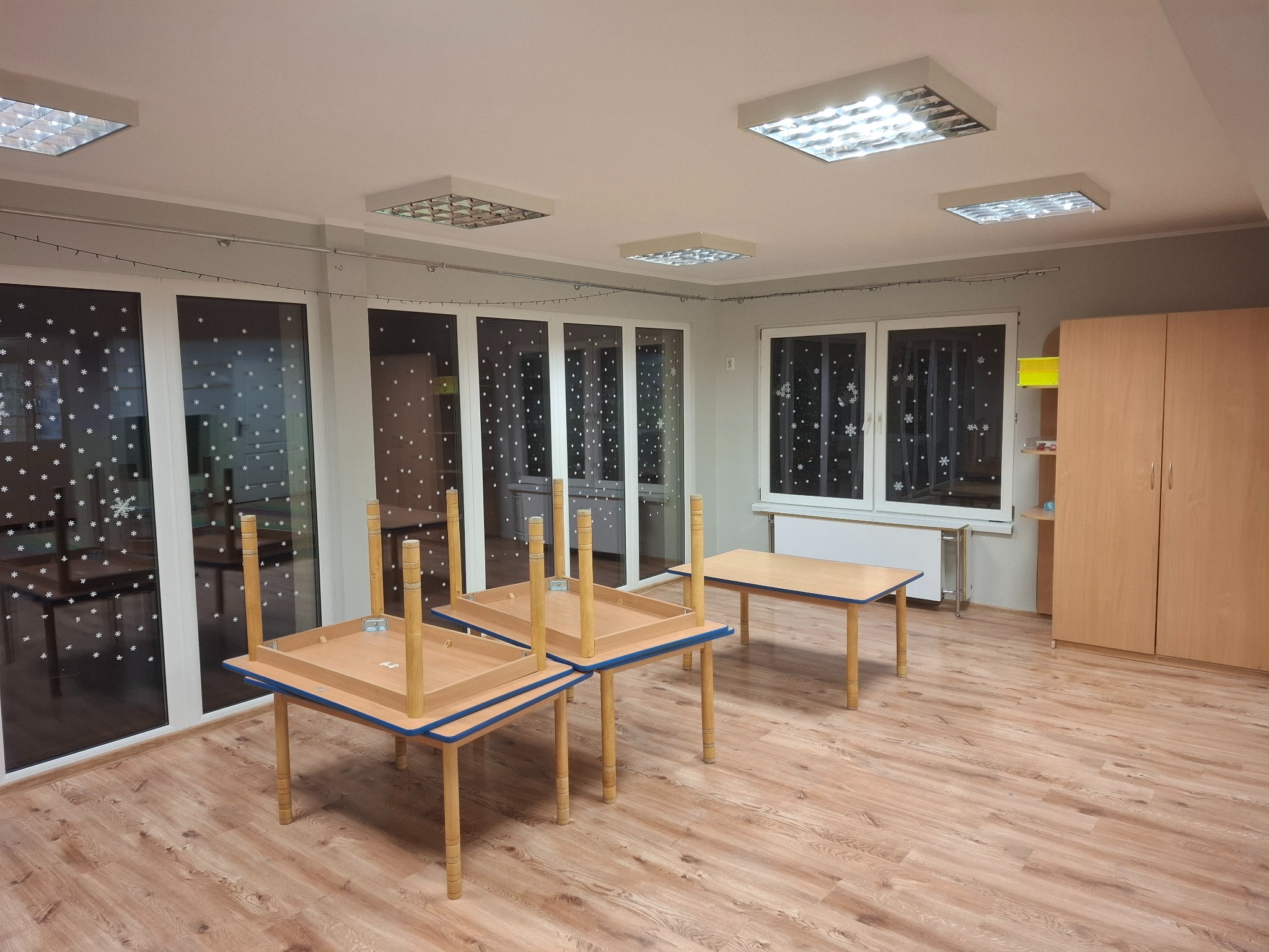 Jasne wnętrze pokoju z drewnianą podłogą, trzema stołami (dwa odwrócone do góry nogami) i szafą. Okna ozdobione śnieżynkami. Oświetlenie sufitowe.