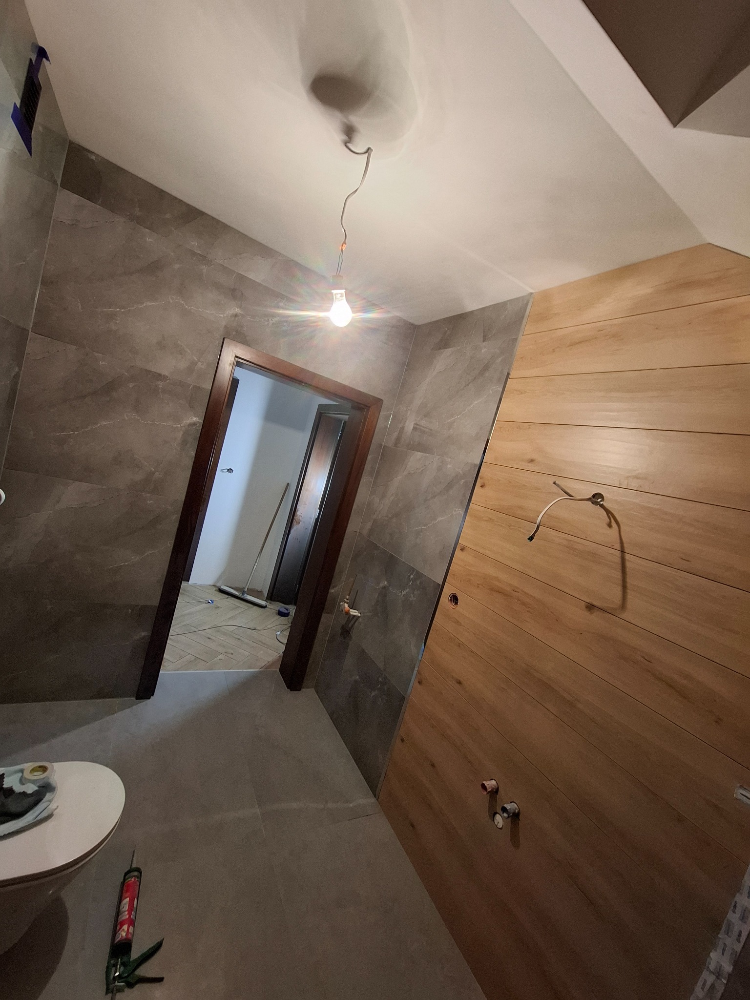 Wykończona łazienka z brązowymi płytkami imitującymi drewno na ścianie i szarymi płytkami imitującymi marmur. Widoczna toaleta i narzędzia glazurnicze.