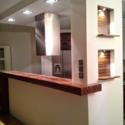 NOVA TECH CZESŁAWA GEMBROWICZ - Nowoczesna kuchnia z drewnianym blatem i wnękami oświetleniowymi. Wykończenie wnętrz w Katowicach. Widoczny fragment podłogi i okap.