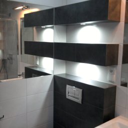 NOVA TECH CZESŁAWA GEMBROWICZ - Nowoczesna łazienka z podświetlanymi półkami z ciemnych płytek, białe płytki na ścianach, lustro i umywalka. Minimalistyczny design.