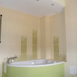 NOVA TECH CZESŁAWA GEMBROWICZ - Jasna łazienka z narożną wanną obudowaną zielonymi płytkami. Na ścianach beżowe płytki z motywem roślinnym. Sufit podwieszany z oświetleniem punktowym.
