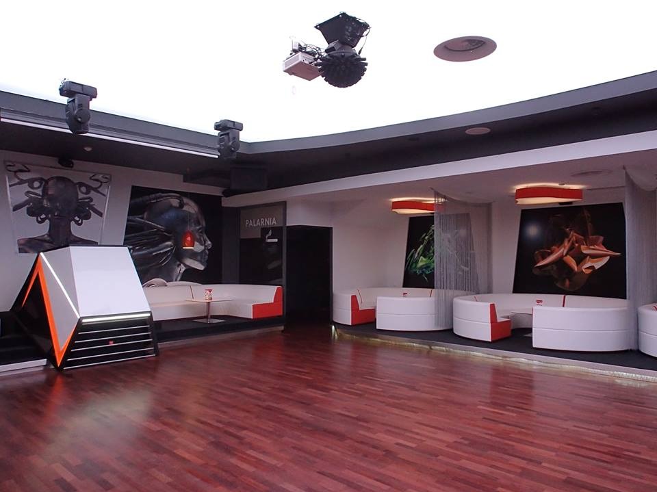 Nowoczesne wnętrze klubu z parkietem, białymi sofami i futurystycznymi dekoracjami. Oświetlenie punktowe i grafiki na ścianach tworzą unikalny klimat.