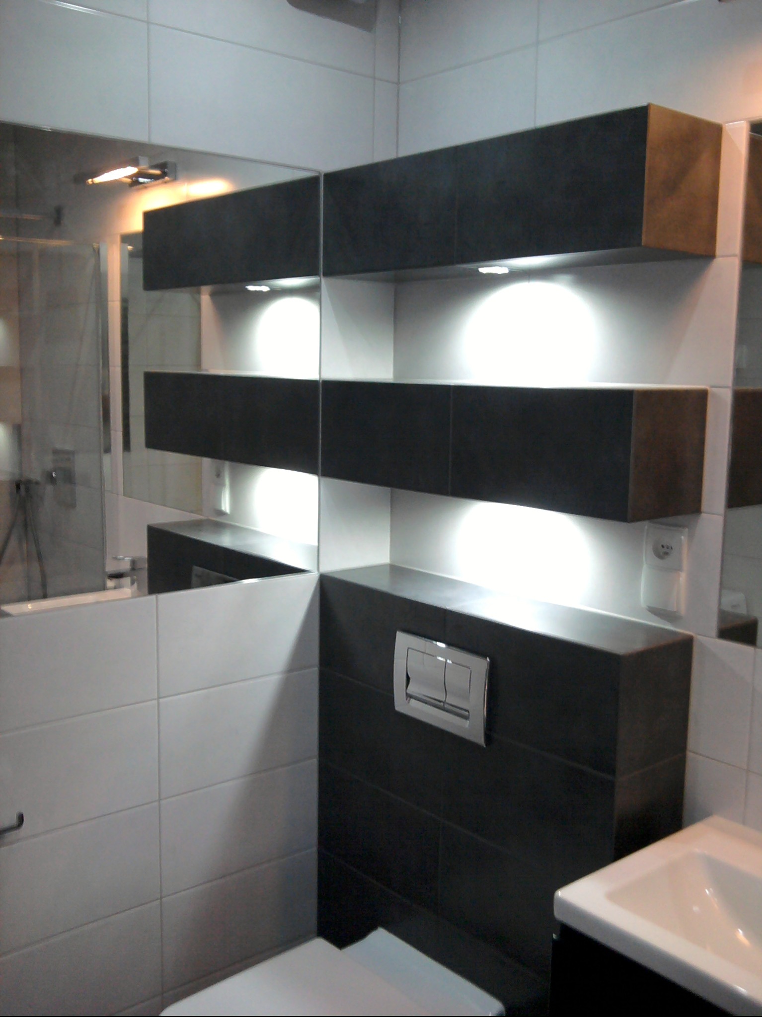 Nowoczesna łazienka z podświetlanymi półkami z ciemnych płytek, białe płytki na ścianach, lustro i umywalka. Minimalistyczny design.