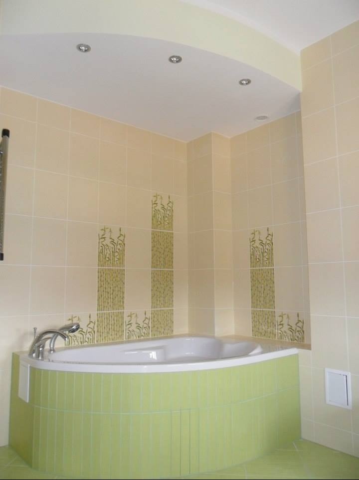 Jasna łazienka z narożną wanną obudowaną zielonymi płytkami. Na ścianach beżowe płytki z motywem roślinnym. Sufit podwieszany z oświetleniem punktowym.
