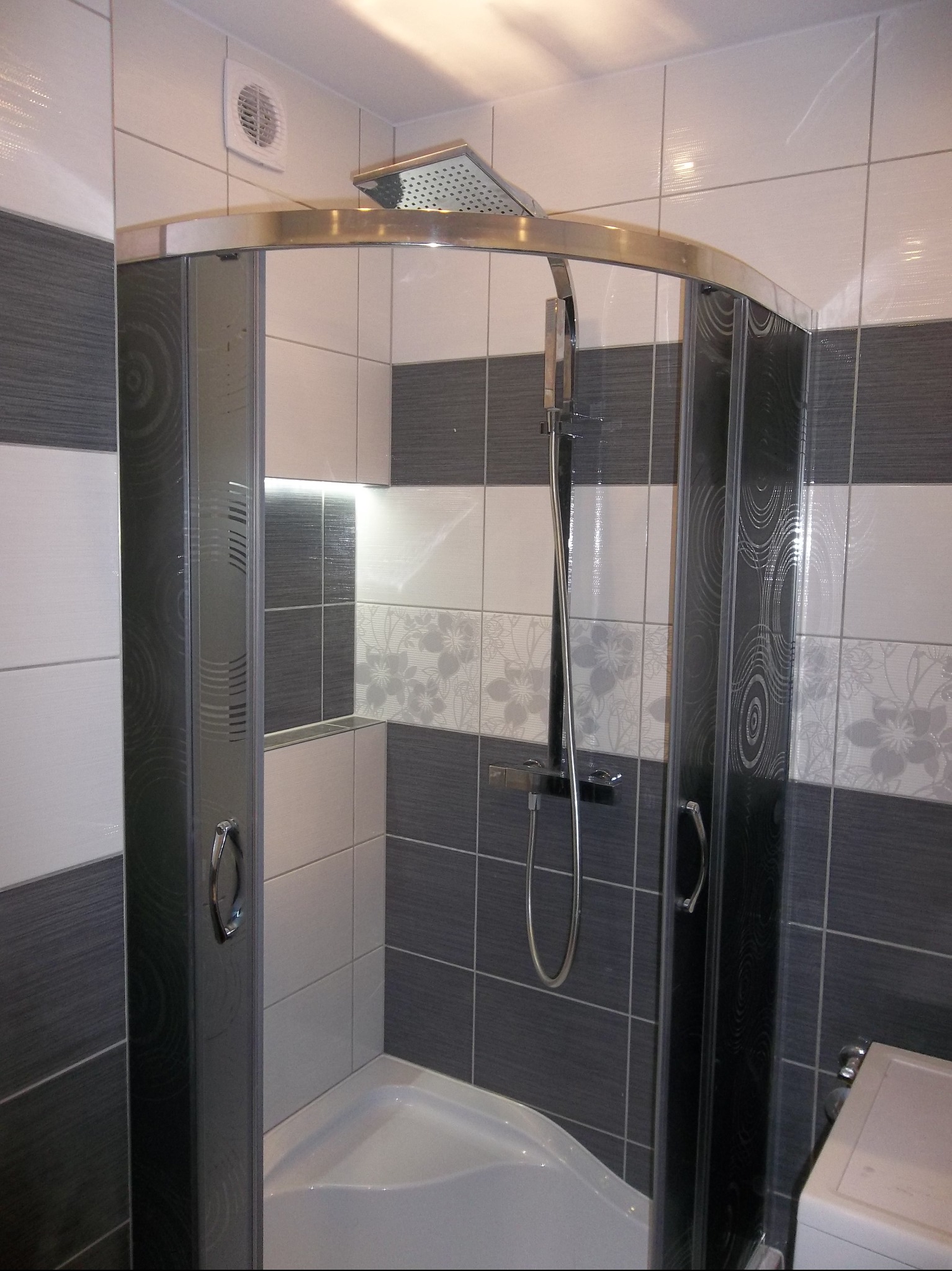 Nowoczesna kabina prysznicowa z chromowanymi profilami i deszczownicą, otoczona płytkami w odcieniach szarości i bieli z subtelnym motywem kwiatowym.