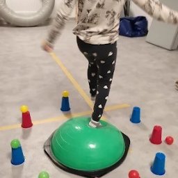 Senso-Help - Małgorzata Pilarska - Fizjoterapia dla dzieci - Dziewczynka ćwiczy równowagę na zielonej półkuli Bosu, otoczonej kolorowymi pachołkami. Sala z oknem i materacami w tle. Ćwiczenia rehabilitacyjne dla dzieci.