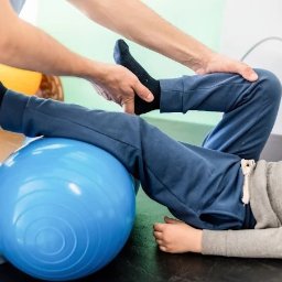 Senso-Help - Małgorzata Pilarska - Fizjoterapia dla dzieci - Rehabilitacja dziecka z użyciem niebieskiej piłki gimnastycznej. Fizjoterapeuta w niebieskim uniformie pomaga w ćwiczeniach, trzymając stopę dziecka.