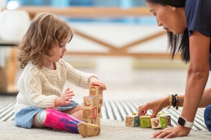 Dziewczynka z protezą nogi układa wieżę z drewnianych klocków, wspierana przez dorosłą osobę. Zabawa edukacyjna w jasnym wnętrzu.