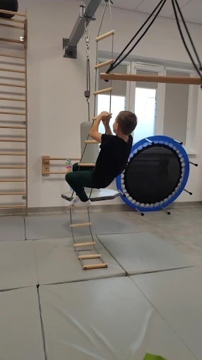 Chłopiec ćwiczy na drabince sznurowej w sali rehabilitacyjnej z materacami, drabinkami gimnastycznymi i trampoliną. Terapia integracji sensorycznej.