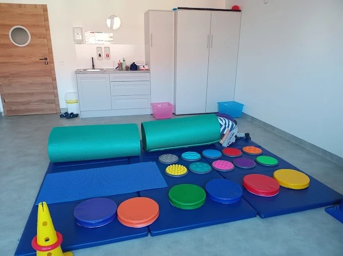 Sala do integracji sensorycznej z tunelem, matą i kolorowymi dyskami. Dziecko w tunelu. Jasne wnętrze, białe meble, drewniane drzwi z okrągłym oknem.