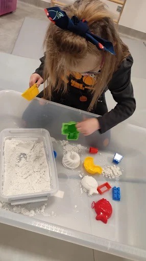 Dziewczynka z kokardą na włosach bawi się w piaskownicy kinetycznej, używając foremek w kształcie rybki i zamku. Terapia sensoryczna w gabinecie.