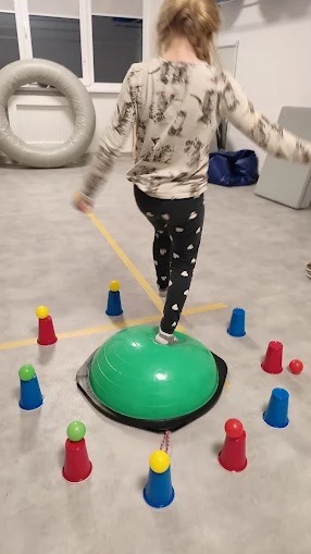 Dziewczynka ćwiczy równowagę na zielonej półkuli Bosu, otoczonej kolorowymi pachołkami. Sala z oknem i materacami w tle. Ćwiczenia rehabilitacyjne dla dzieci.