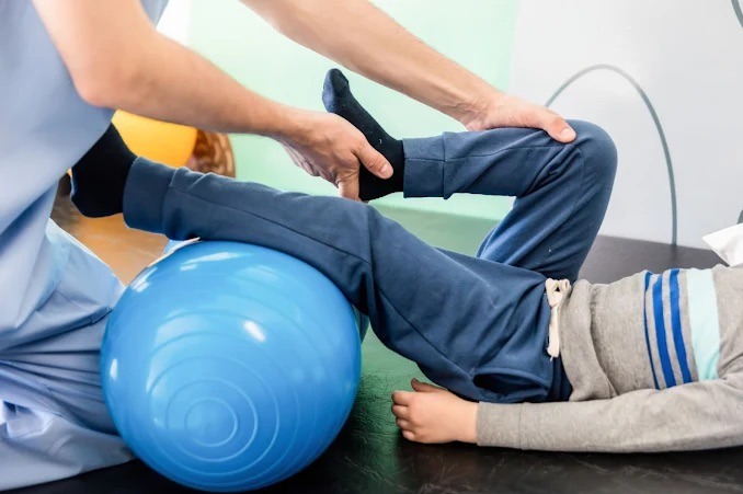 Rehabilitacja dziecka z użyciem niebieskiej piłki gimnastycznej. Fizjoterapeuta w niebieskim uniformie pomaga w ćwiczeniach, trzymając stopę dziecka.