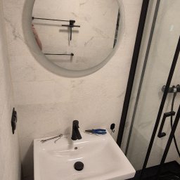 WOŹNICA USŁUGI BUDOWLANO-REMONTOWE SPÓŁKA Z OGRANICZONĄ ODPOWIEDZIALNOŚCIĄ - Nowoczesna łazienka po remoncie: biała umywalka z czarną armaturą, okrągłe lustro z podświetleniem i czarna kabina prysznicowa. Widoczne narzędzia glazurnicze.