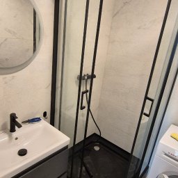 WOŹNICA USŁUGI BUDOWLANO-REMONTOWE SPÓŁKA Z OGRANICZONĄ ODPOWIEDZIALNOŚCIĄ - Nowoczesna łazienka po remoncie: czarna kabina prysznicowa, umywalka z szafką w odcieniach szarości, okrągłe lustro i pralka. Marmurowe płytki na ścianach i podłodze.