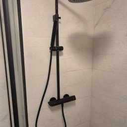 WOŹNICA USŁUGI BUDOWLANO-REMONTOWE SPÓŁKA Z OGRANICZONĄ ODPOWIEDZIALNOŚCIĄ - Nowoczesna kabina prysznicowa z czarną armaturą i deszczownicą na tle jasnych, marmurkowych płytek. Minimalistyczny design, widoczny fragment odpływu.