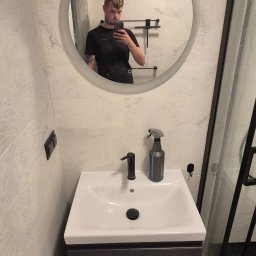 WOŹNICA USŁUGI BUDOWLANO-REMONTOWE SPÓŁKA Z OGRANICZONĄ ODPOWIEDZIALNOŚCIĄ - Nowoczesna łazienka z okrągłym lustrem, białą umywalką z szarą szafką i kabiną prysznicową z czarnymi ramami. Widoczne płytki imitujące marmur.