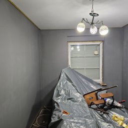 WOŹNICA USŁUGI BUDOWLANO-REMONTOWE SPÓŁKA Z OGRANICZONĄ ODPOWIEDZIALNOŚCIĄ - Pokój po malowaniu na szaro, zabezpieczony folią i krzesłem. Widoczna lampa sufitowa i okno. Świeżo pomalowane ściany z żółtą listwą pod sufitem.
