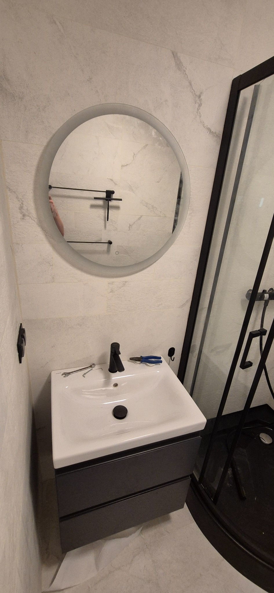 Nowoczesna łazienka po remoncie: biała umywalka z czarną armaturą, okrągłe lustro z podświetleniem i czarna kabina prysznicowa. Widoczne narzędzia glazurnicze.