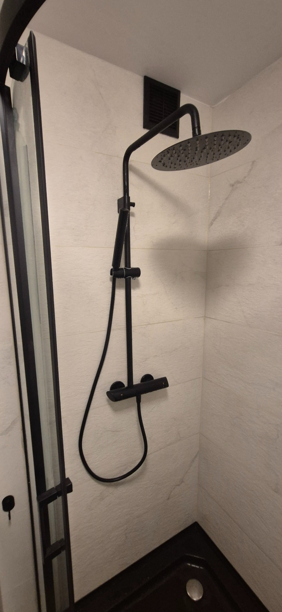 Nowoczesna kabina prysznicowa z czarną armaturą i deszczownicą na tle jasnych, marmurkowych płytek. Minimalistyczny design, widoczny fragment odpływu.